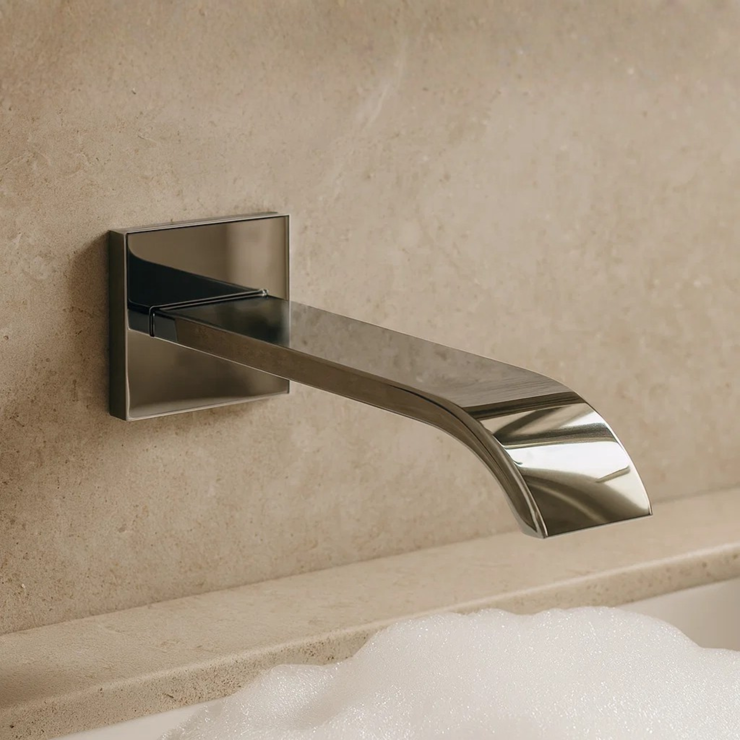 Chrome wall-mounted faucet on a beige tiled wall with a bathtub in the foreground.